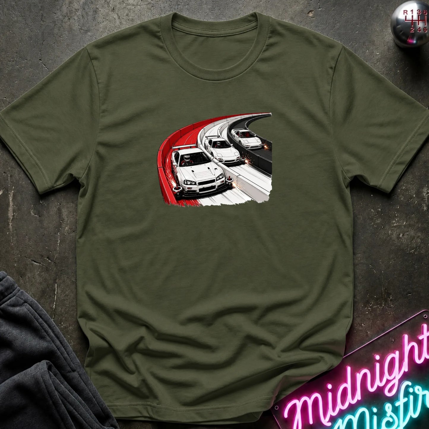An olive green crewneck t-shirt featuring a graphic print of three white sports cars racing on a curve, styled flat-lay on a dark textured surface with a neon sign and dark pants in the background.