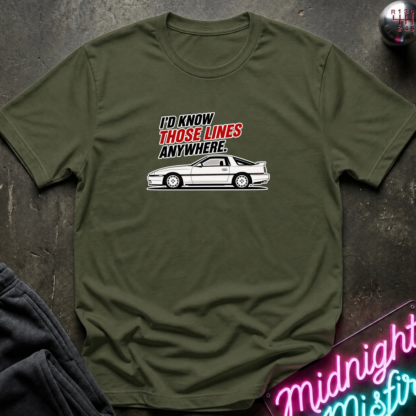An olive green short-sleeve t-shirt with a graphic print of a white sports car and the text 'I'D KNOW THOSE LINES ANYWHERE.' in black and red, laid flat on a dark textured surface.
