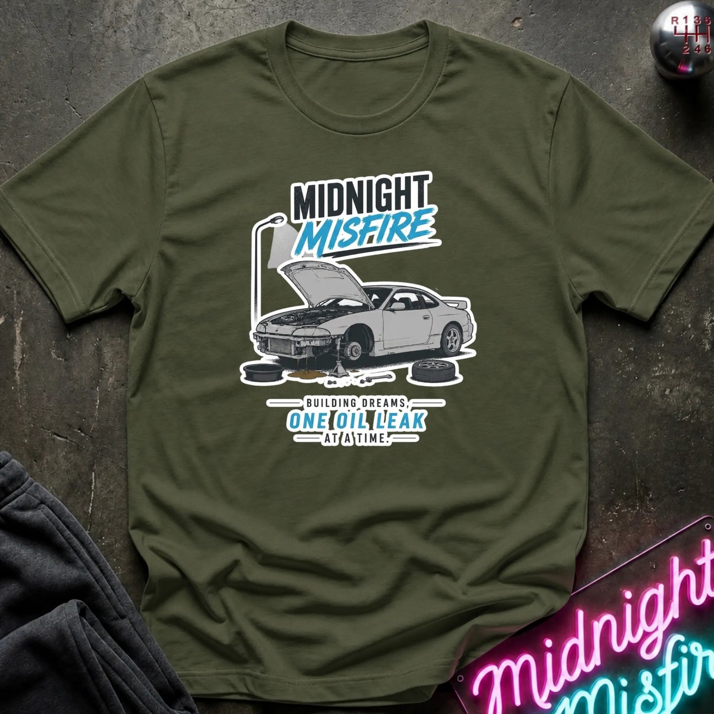 An olive green graphic t-shirt laid flat on a dark textured surface. The shirt features a white and blue graphic of a car with its hood open, surrounded by oil cans, with the text 'MIDNIGHT MISFIRE' and 'BUILDING DREAMS ONE OIL LEAK AT ANY TIME'.