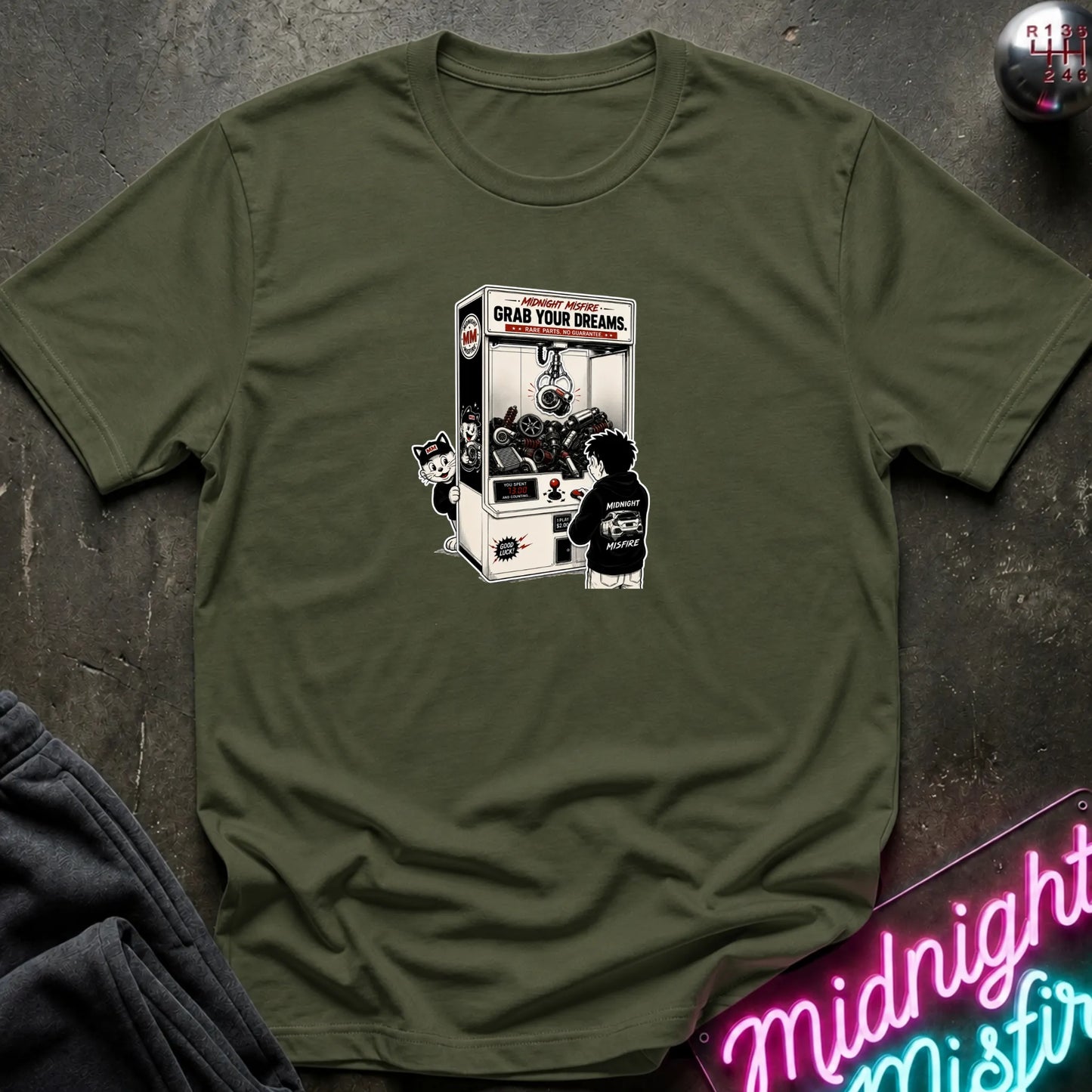 An olive green short-sleeve crewneck t-shirt laid flat on a dark textured surface, featuring a central graphic of a person in a black hoodie playing a claw machine. 