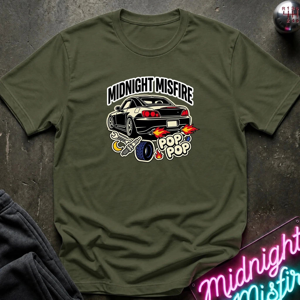 An olive green short-sleeve t-shirt with a graphic of a black sports car and the text 'MIDNIGHT MISFIRE' and 'POP POP' with flames, laid flat on a dark textured surface.