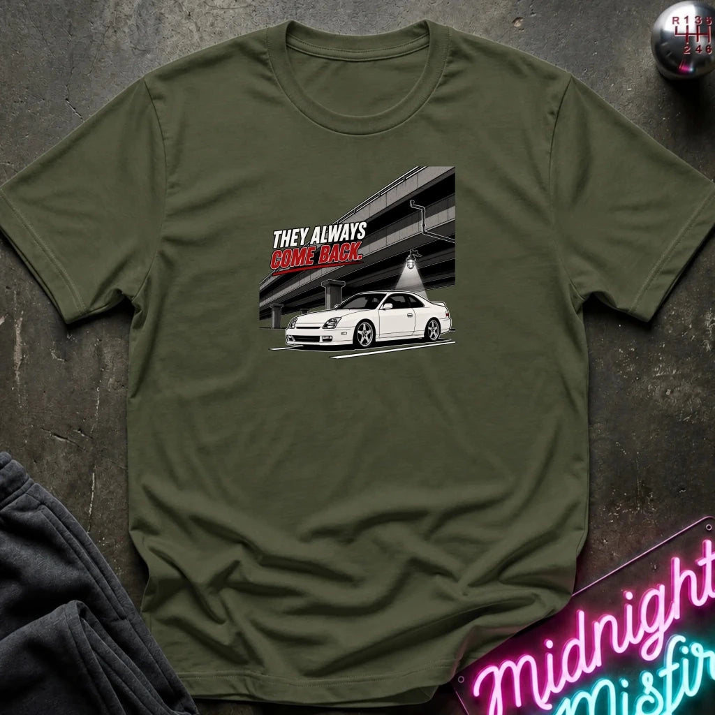 An olive green short-sleeve t-shirt laid flat on a dark textured surface, featuring a graphic print of a white sports car under an overpass 