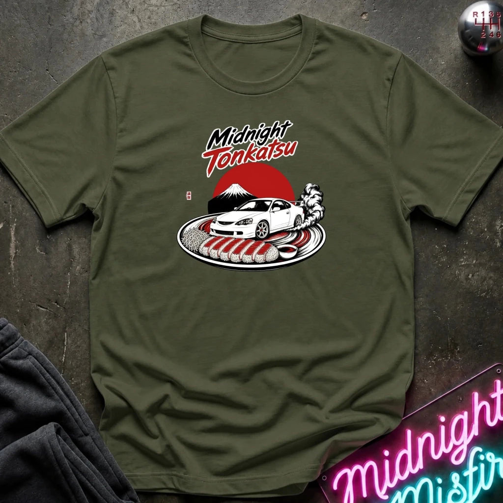 An olive green crewneck t-shirt featuring a 'Midnight Tenkatsu' graphic print on the chest, styled flat on a dark textured surface with a neon sign and dark pants partially visible.