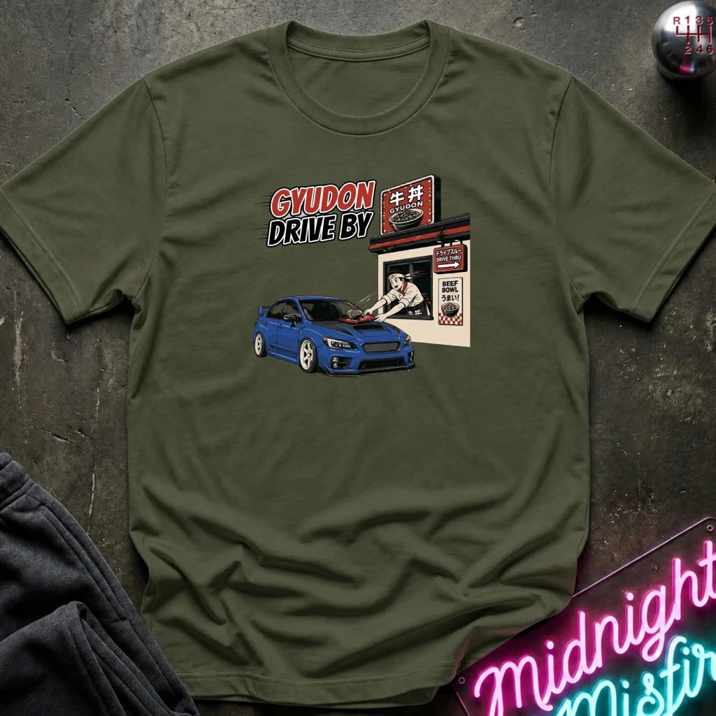 An olive green graphic t-shirt laid flat on a dark textured surface. The graphic features a blue car and a drive-by restaurant scene with the text 'GYUDON DRIVE BY'.