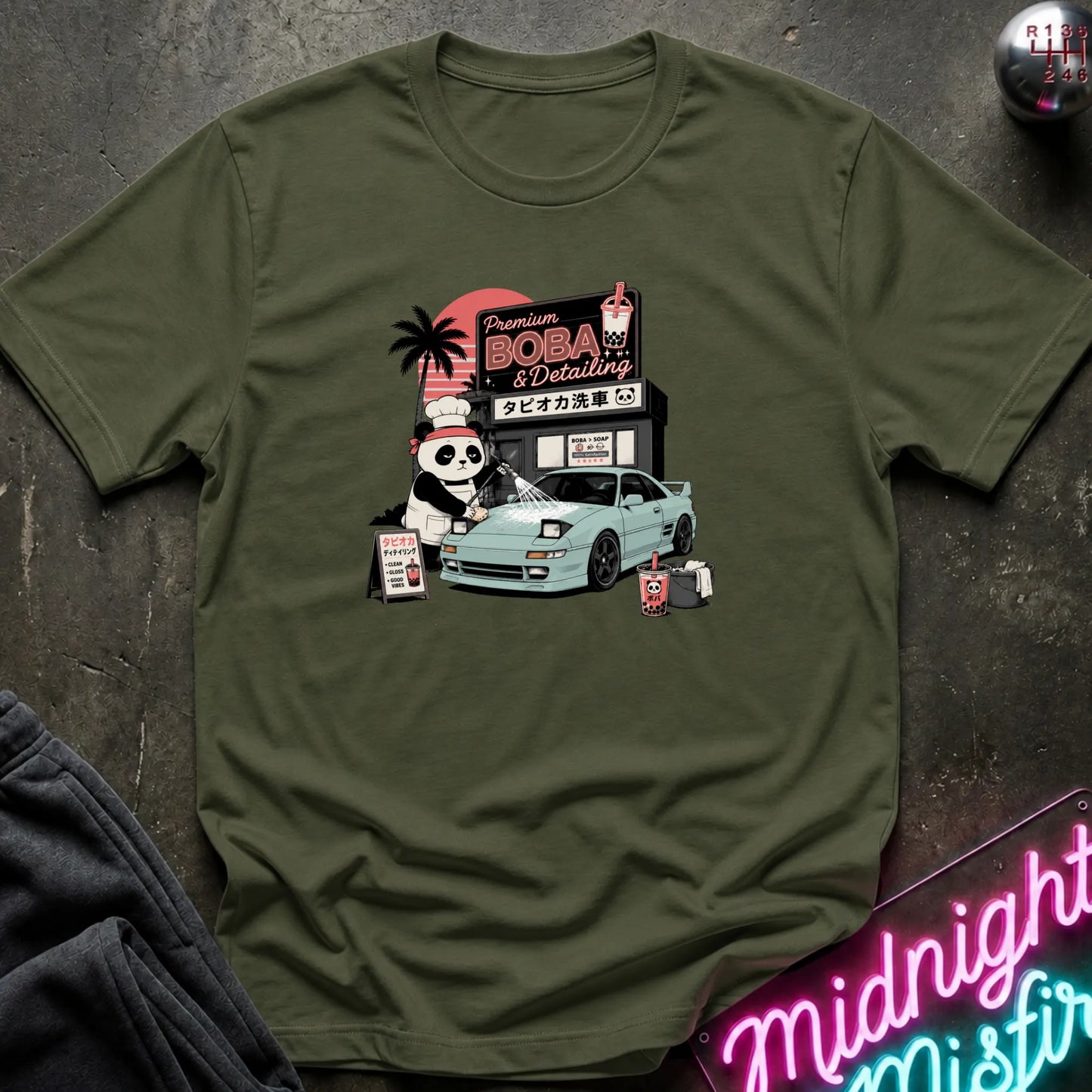 An olive green short-sleeve graphic T-shirt laid flat on a dark textured surface. The shirt features a colorful retro-style print of a panda character, a light blue sports car, and a sign reading 'Premium BOBA & Coffee' with Japanese text.