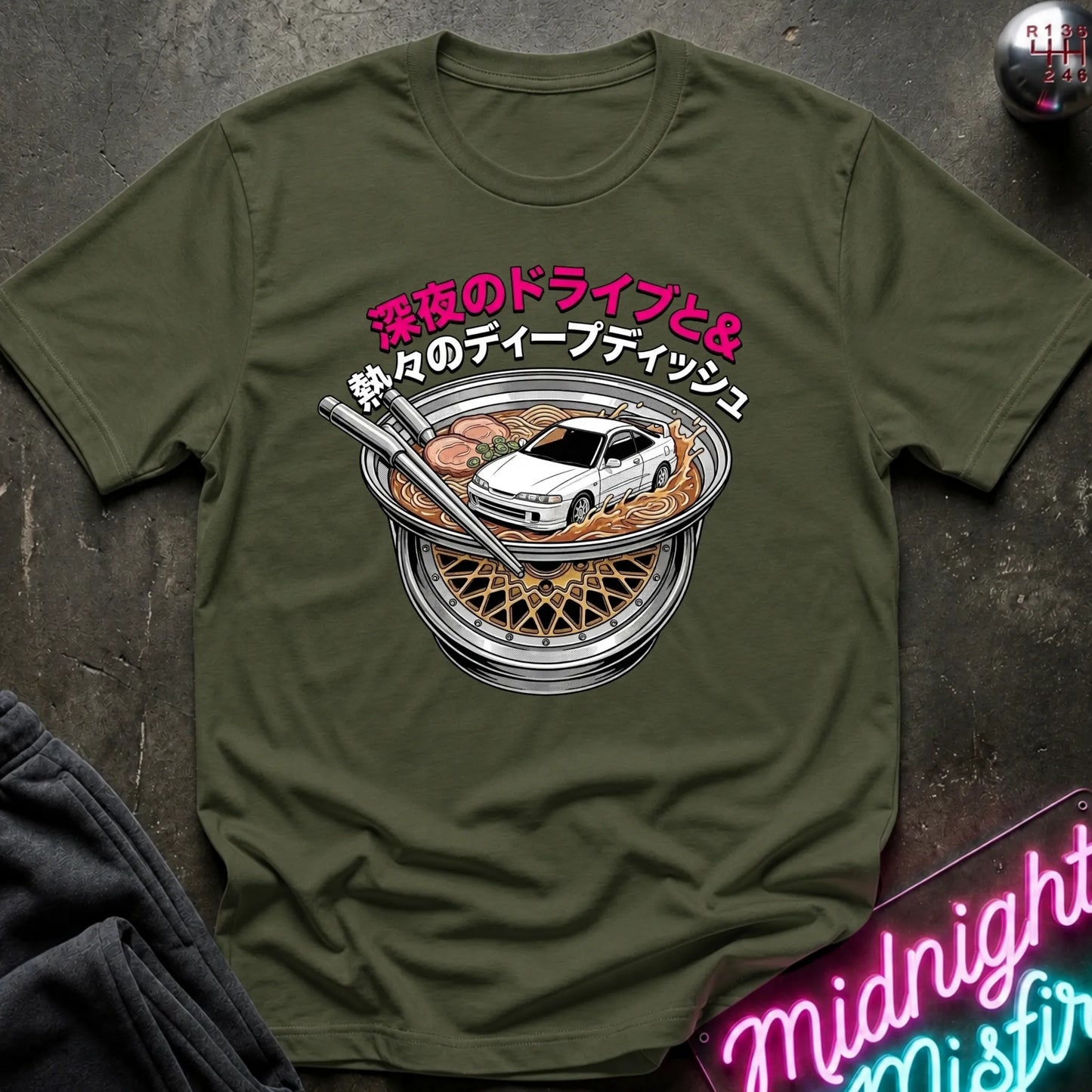 An olive green short-sleeve t-shirt with a graphic print of a white sports car inside a bowl of ramen, featuring pink and white Japanese text. The shirt is laid flat on a dark textured surface with a neon sign and dark fabric partially visible in the corners.