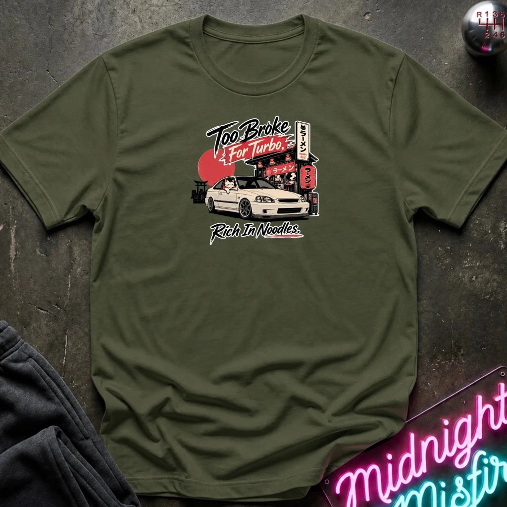 An olive green short-sleeve t-shirt with a graphic print of a white car and Japanese-style signage, laid flat on a dark textured surface with a neon sign and dark pants partially visible.