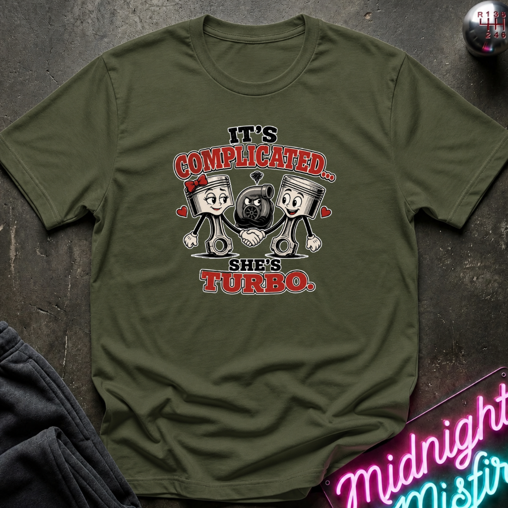 An olive green graphic t-shirt laid flat on a dark textured surface. The graphic features two cartoon piston characters with a figure in the middle, with the text 'IT'S COMPLICATED.