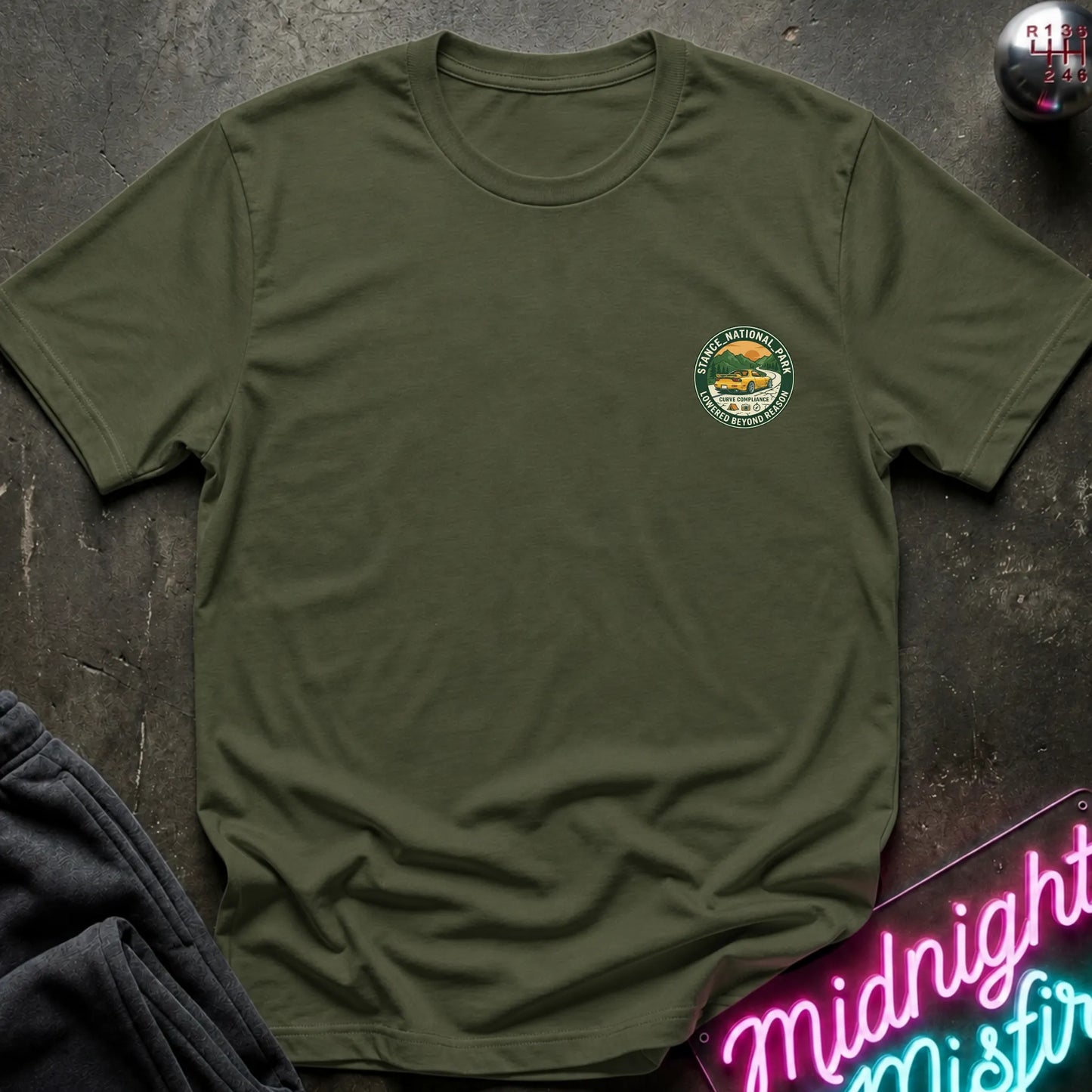An olive green crewneck t-shirt with a circular logo on the left chest, laid flat on a dark textured surface with a neon sign and dark pants in the background.