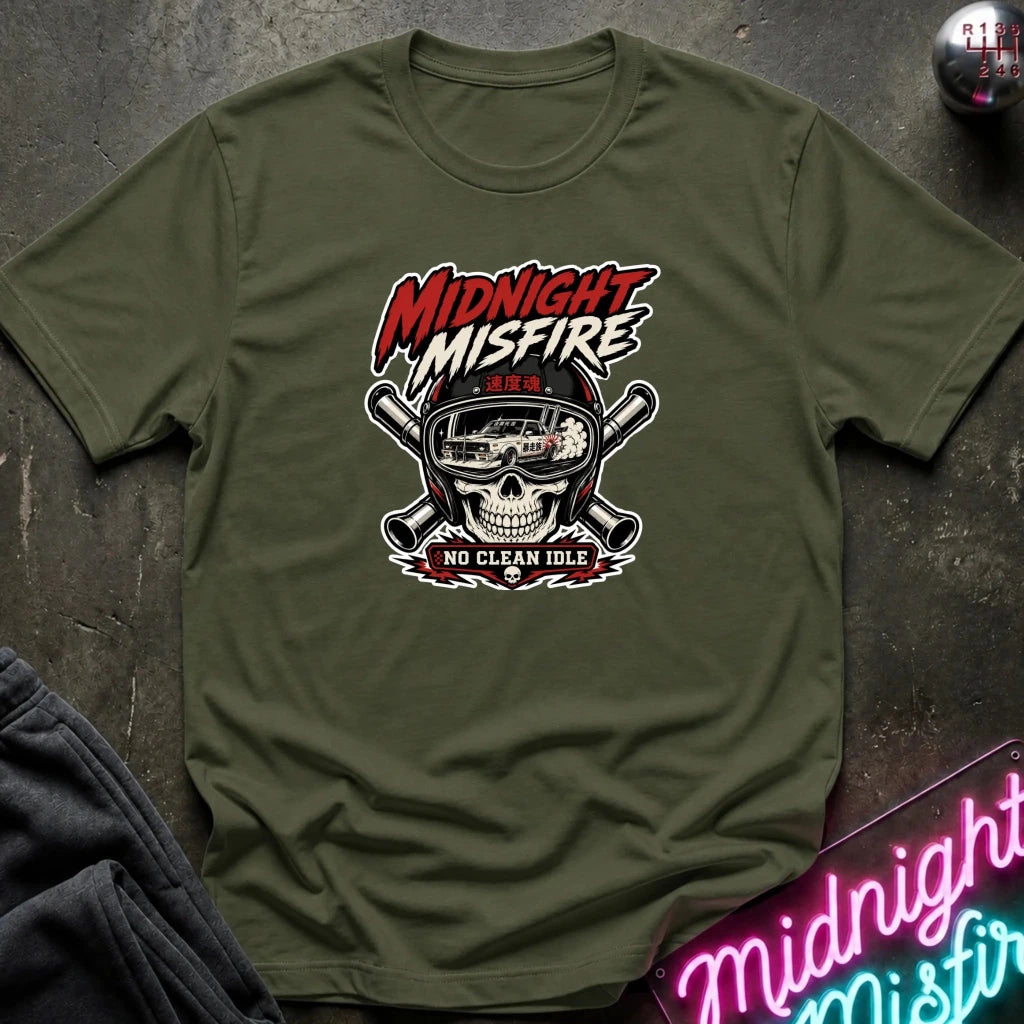 An olive green graphic t-shirt laid flat on a dark textured surface. The shirt features a central graphic of a skull wearing a racing helmet with crossed pipes behind it, and the text 'MIDNIGHT MISFIRE' and 'NO CLEAN IDLE'.