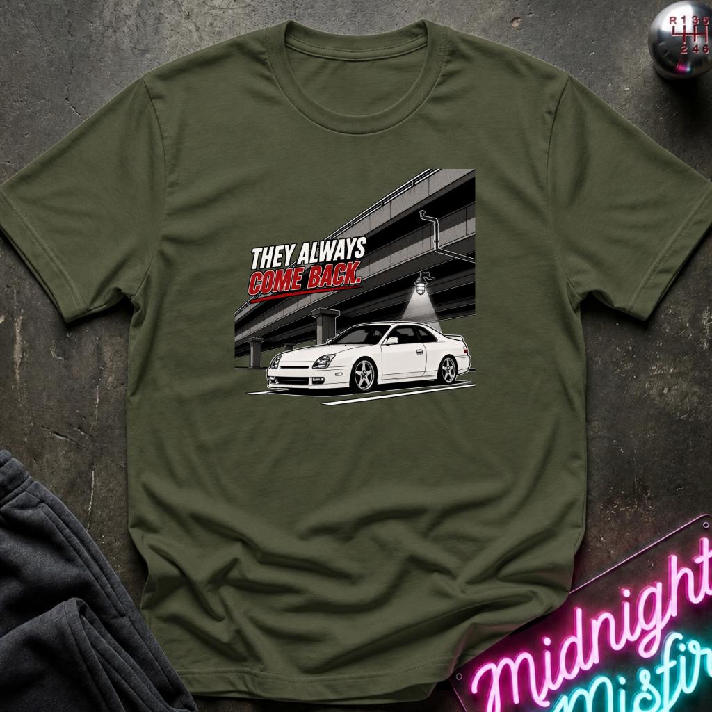 An olive green short-sleeve t-shirt with a graphic print of a white sports car and the text 'THEY ALWAYS COME BACK' in white and red, laid flat on a dark textured surface with decorative props.