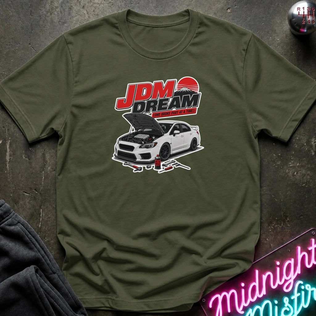 An olive green crewneck t-shirt laid flat on a dark textured surface, featuring a graphic of a white sports car with its hood open beneath the red and black 'JDM DREAM' text.