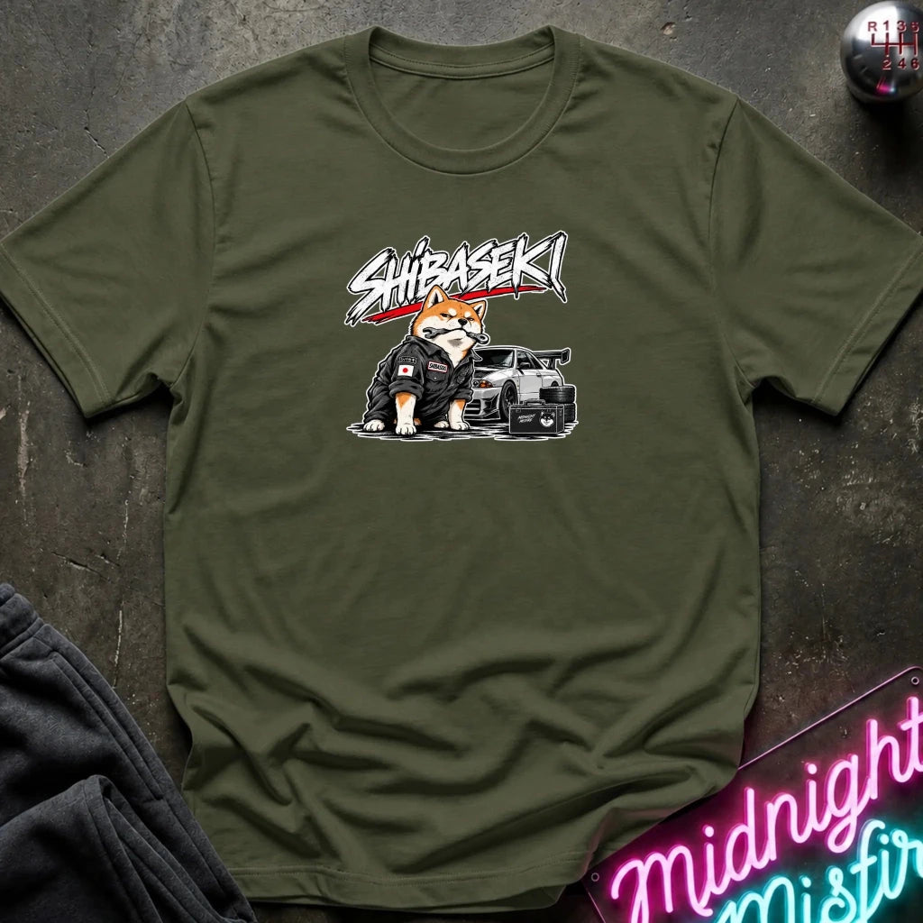 An olive green crewneck t-shirt featuring a graphic of a Shiba Inu dog in a black jacket next to a black sports car, with the text 'SHIBASEKI' printed above. The shirt is laid flat on a dark textured surface with lifestyle props including a neon sign and folded sweatpants.