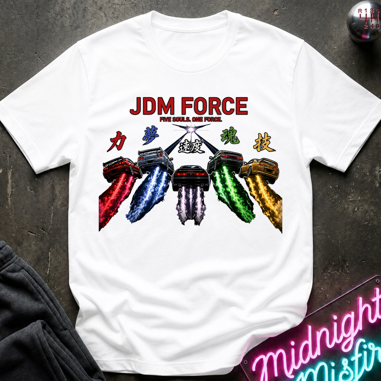 A white short-sleeve graphic T-shirt laid flat on a dark textured surface. The shirt features a colorful chest print with the words 'JDM FORCE' and multiple stylized cars in red, blue, white, green, and gold.