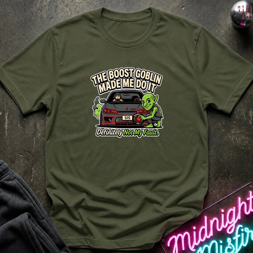 An olive green short-sleeve t-shirt with a graphic print of a green goblin next to a black sports car and the text 'THE BOOST GOBLIN MADE ME DO IT' and 'Definitely Not My Fault'. The shirt is laid flat on a dark textured surface with a neon sign and dark fabric partially visible in the corners.