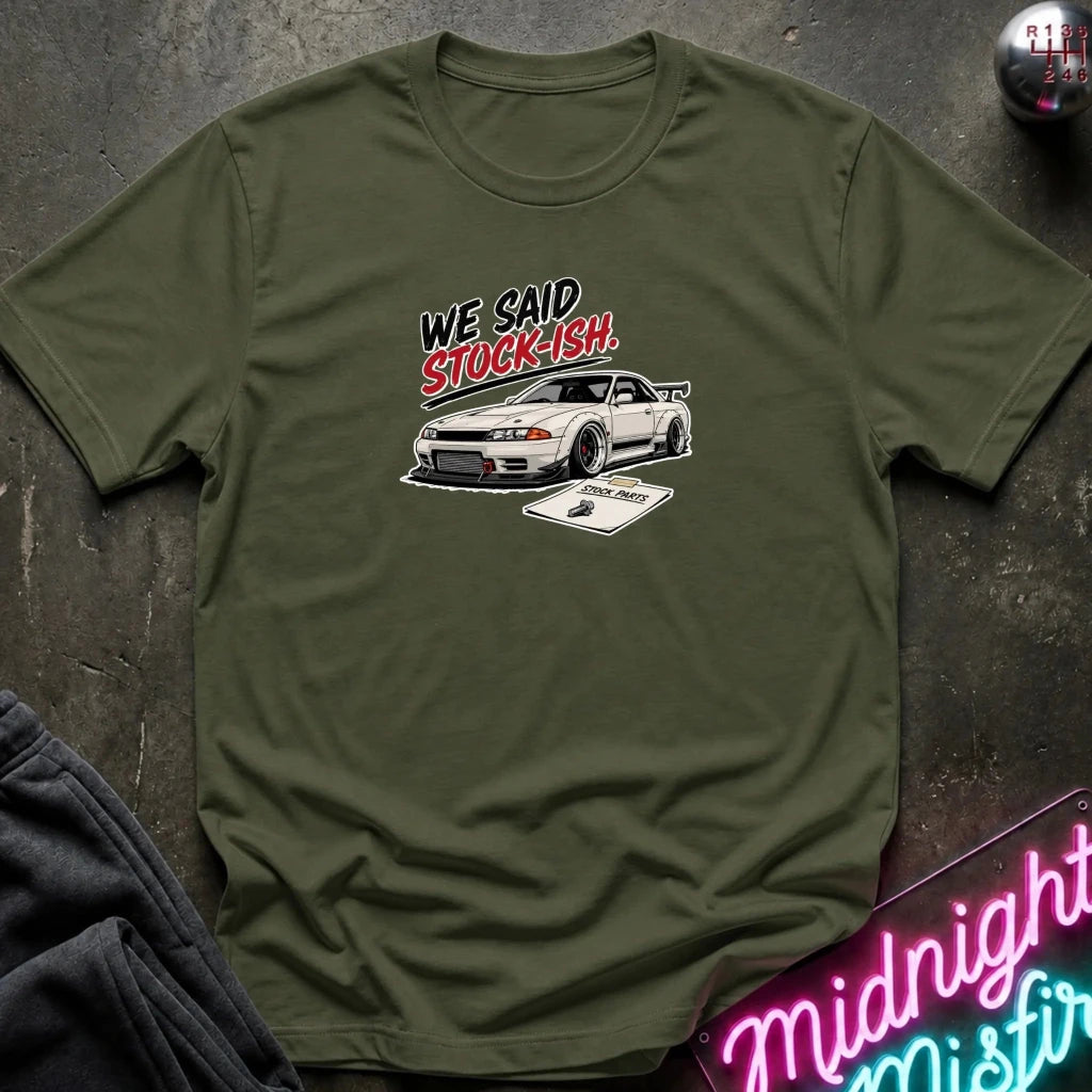 An olive green short-sleeve t-shirt laid flat on a dark textured surface. The shirt features a graphic print of a white sports car with the text 'WE SAID STOCK-ISH.
