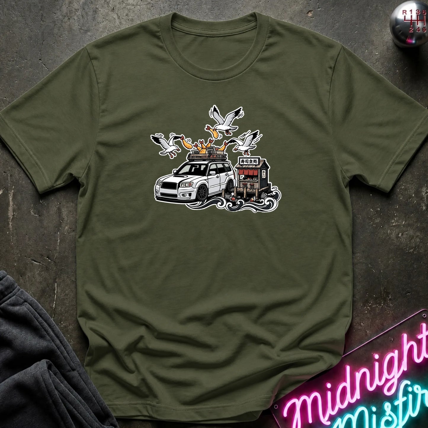 An olive green crewneck t-shirt with a graphic print of a white station wagon with a roof rack, surrounded by seagulls and a small building, laid flat on a dark textured surface.