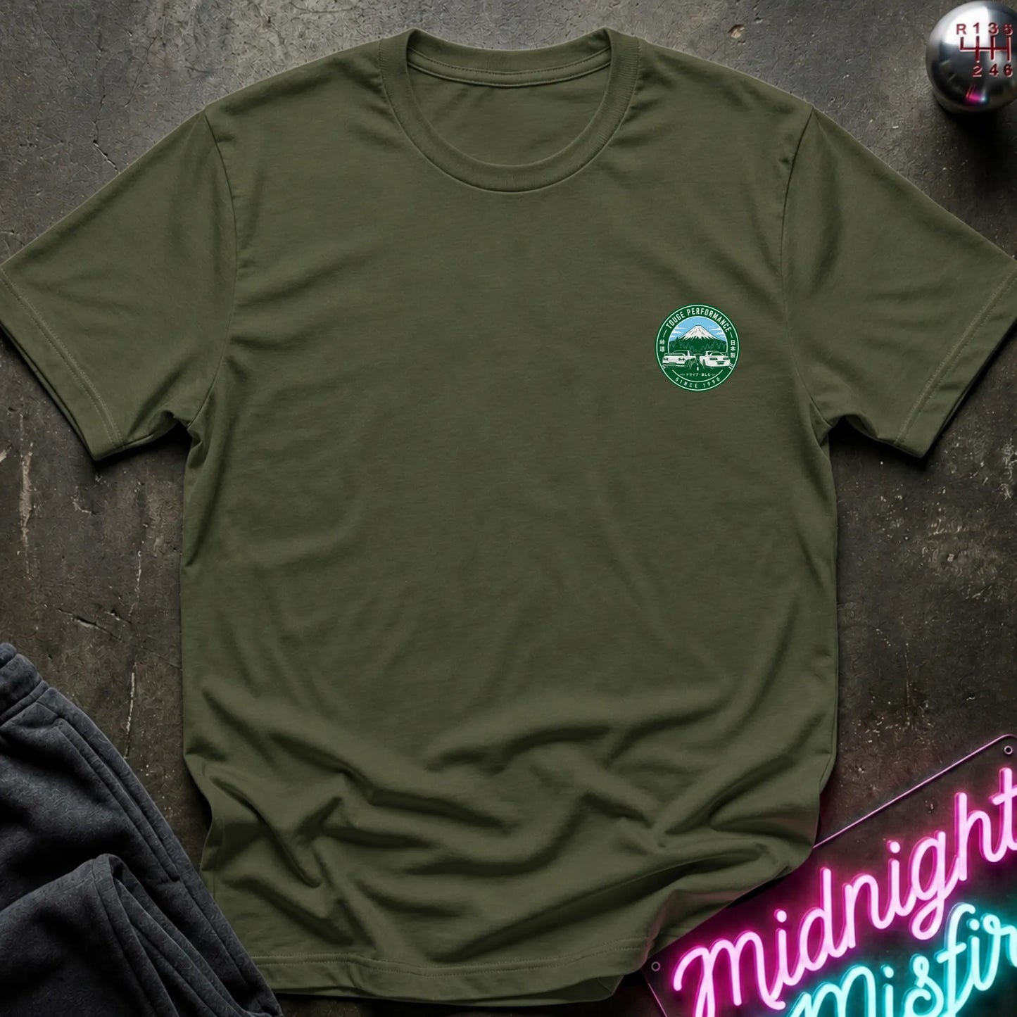 Olive green short-sleeve graphic T-shirt laid flat on a dark textured surface. The shirt features a small circular green and white print logo  with a mountain, two cars, and the text 'TOUGE PERFORMANCE' and 'SINCE 1990'.