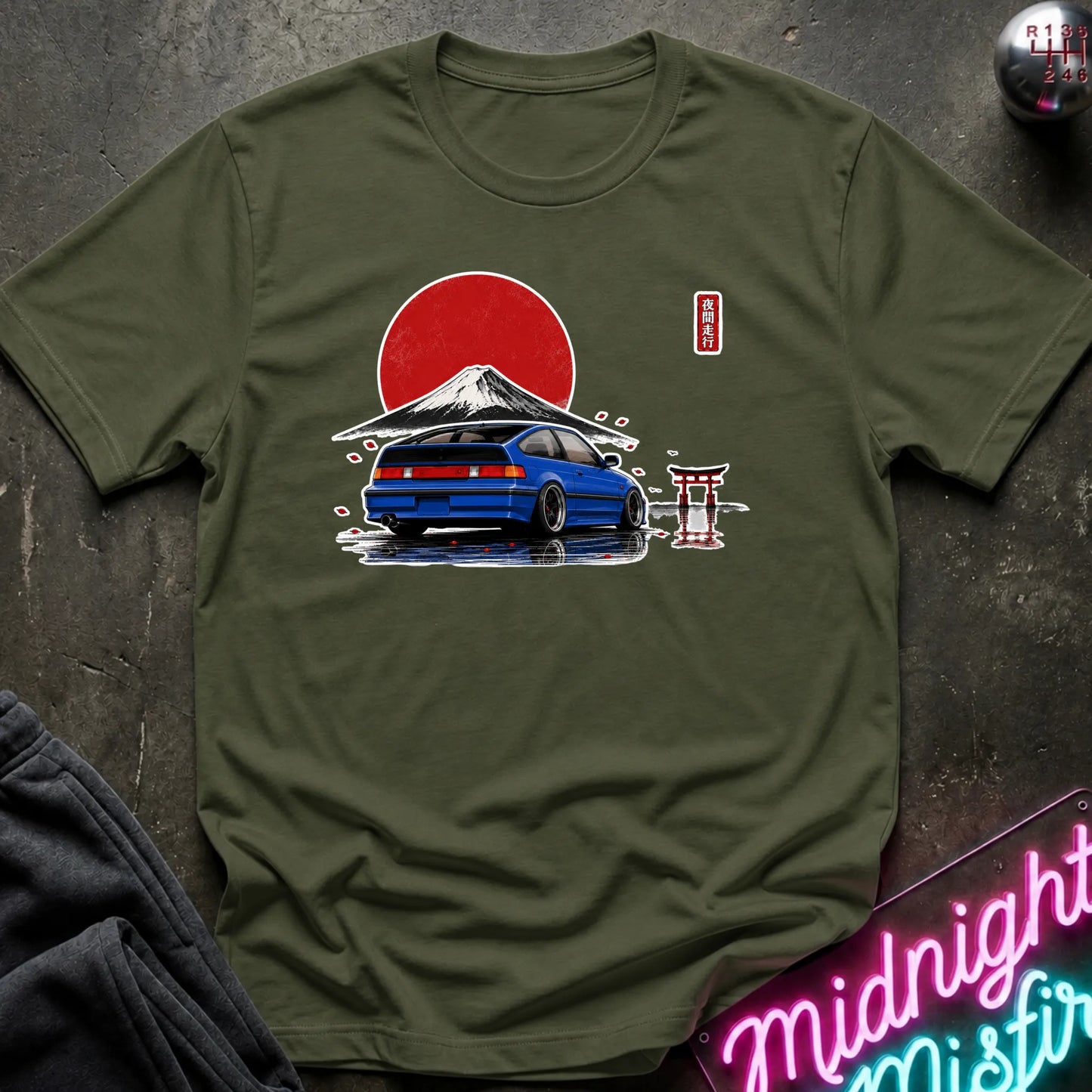 An olive green crewneck t-shirt featuring a graphic print of a blue sports car parked in front of a red sun and Mount Fuji, styled flat-lay on a dark textured surface with a neon sign and dark pants in the background.