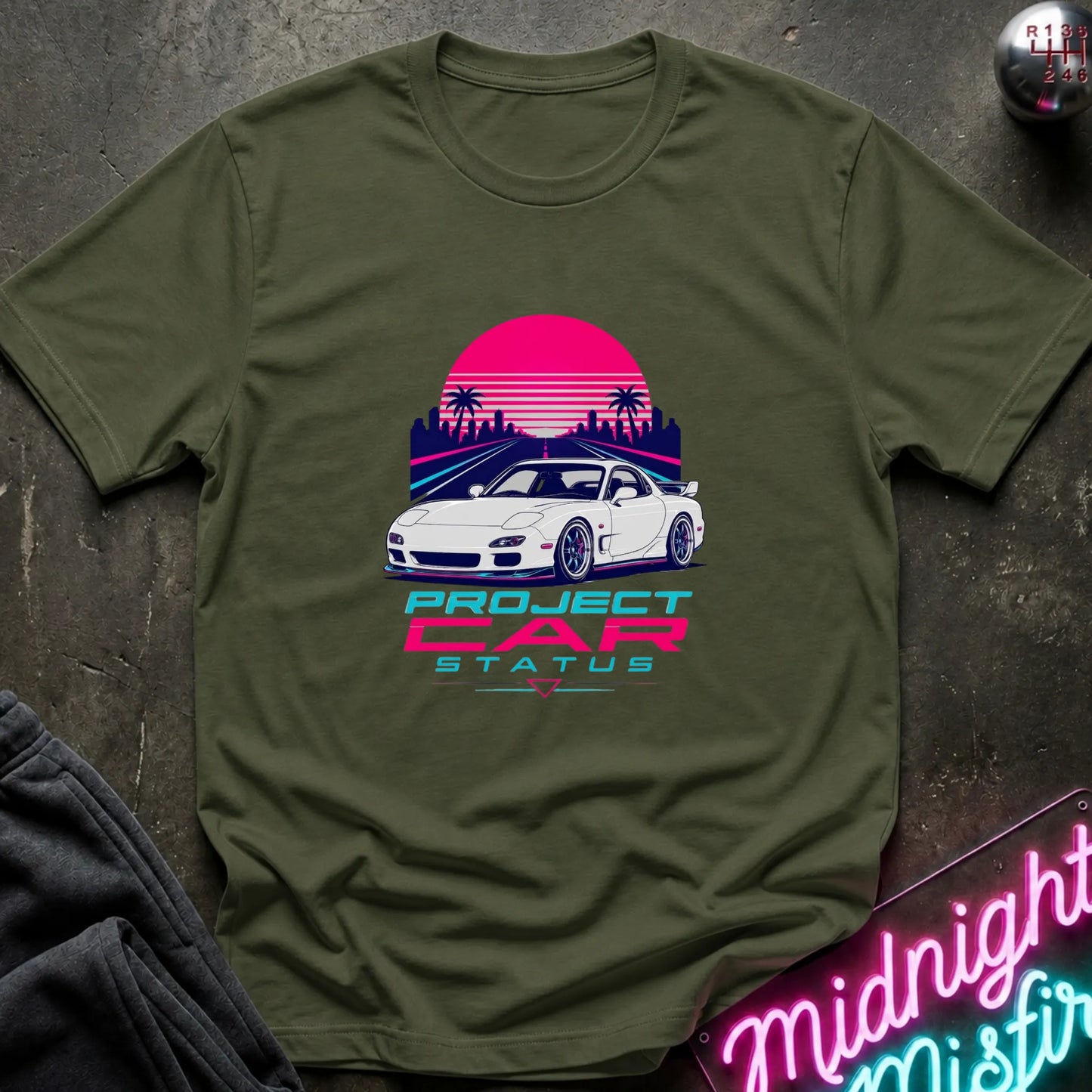 An olive green short-sleeve graphic T-shirt laid flat on a dark textured surface. The shirt features a retro-style print of a white sports car in front of a pink and purple sunset with palm trees, and the text 'PROJECT CAR STATUS' below.