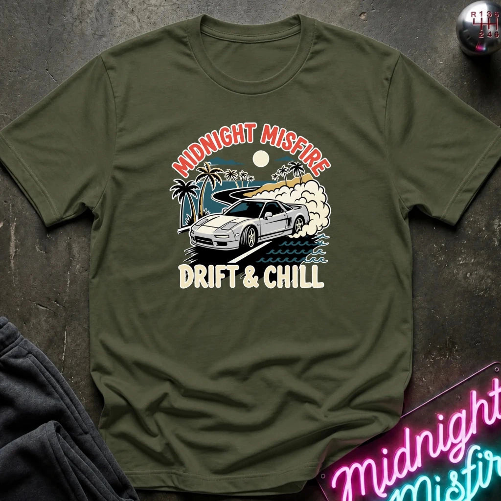 An olive green short-sleeve t-shirt with a retro graphic of a white sports car drifting, palm trees, and a surfboard under the text 'MIDNIGHT MISFIRE DRIFT & CHILL'. The shirt is displayed flat on a dark textured surface with a neon sign and dark pants partially visible.