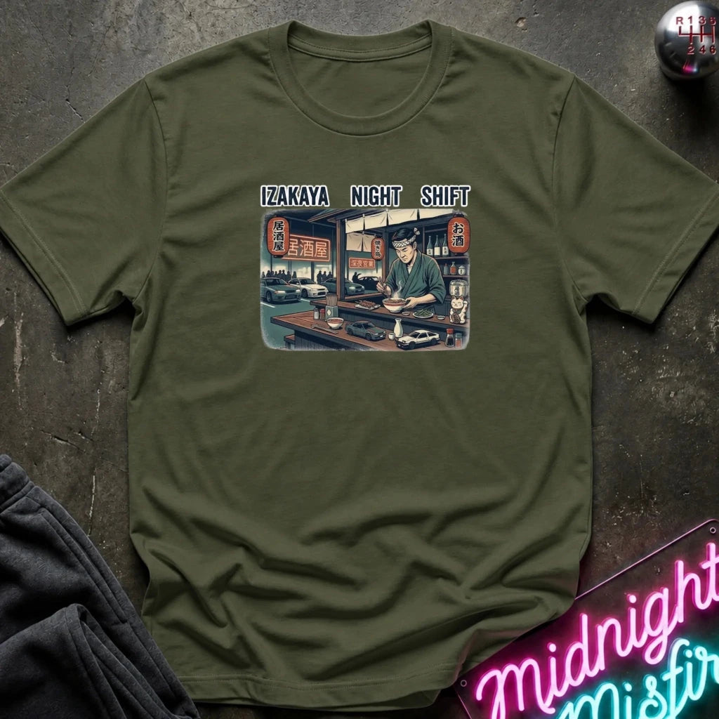An olive green short-sleeve t-shirt with a graphic print on the front reading 'IZAKAYA NIGHT SHIFT' above an illustration of a man working at an izakaya. The shirt is laid flat on a dark textured surface with a neon sign and dark pants partially visible as props.