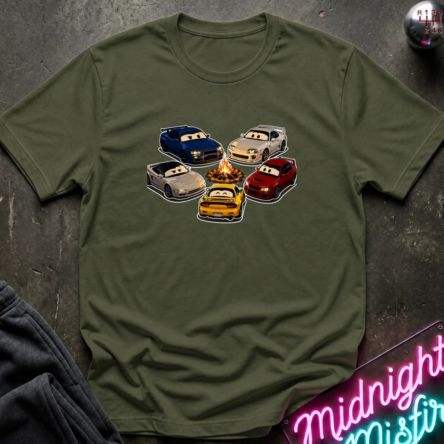 An olive green short-sleeve t-shirt laid flat on a dark textured surface, featuring a graphic of cartoon race cars arranged around a campfire. The shirt is styled with a gray sweatshirt in the corner and a pink neon sign in the bottom right.