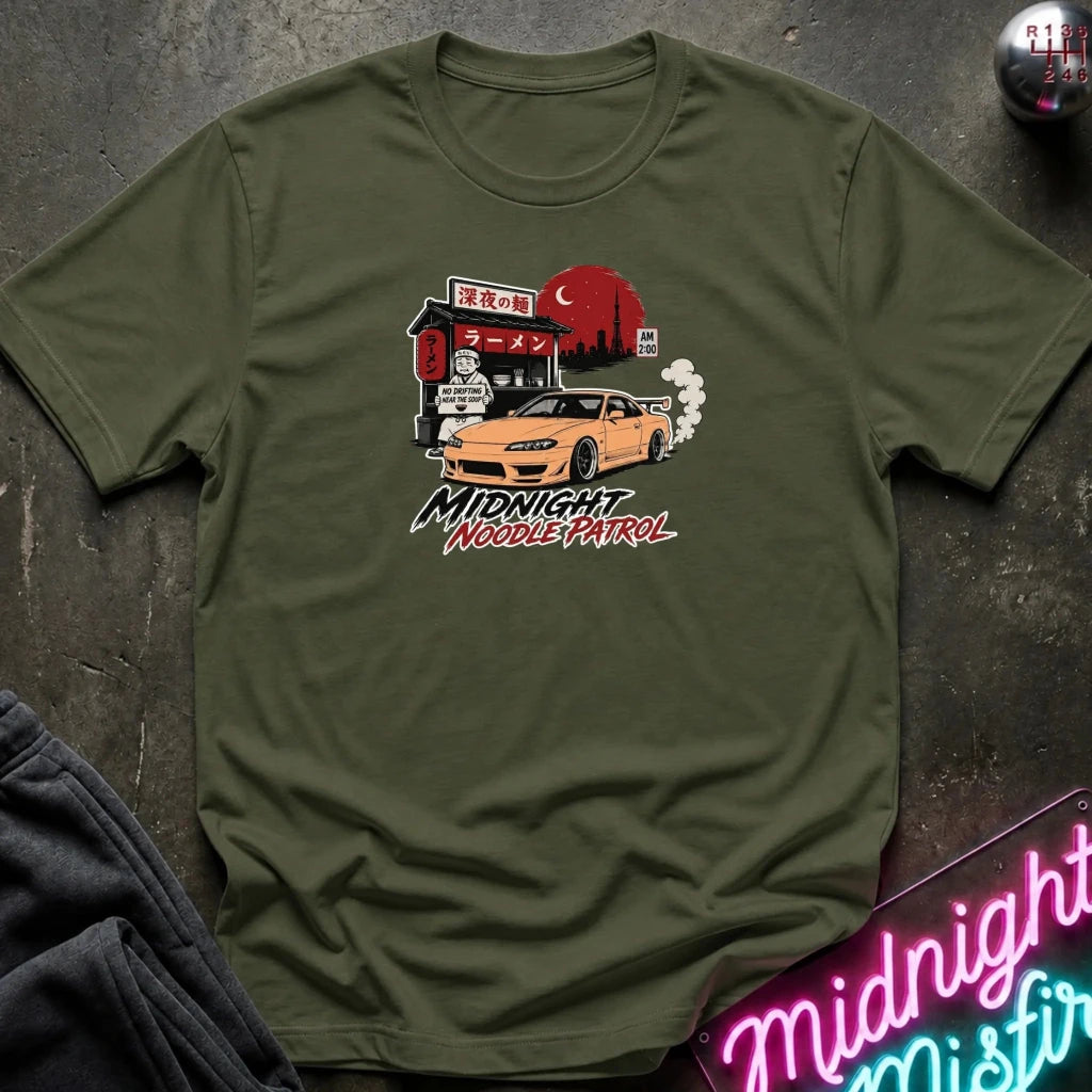 olive crewneck t-shirt featuring a graphic print of a yellow Nissan Silvia S15 drift car in front of a red moon and Japanese storefront, with the text 'MIDNIGHT Noodle PATROL'. The shirt is laid flat on a dark textured surface with a neon sign and dark pants partially visible.