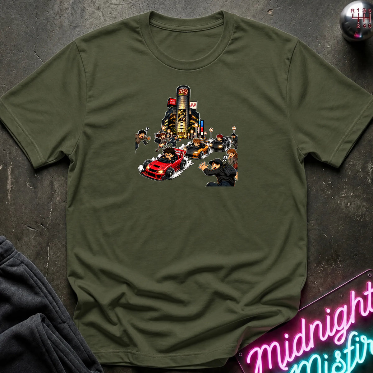 An olive short-sleeve crewneck t-shirt with a graphic print of a city street scene featuring a red sports car and small figures, laid flat on a dark textured surface with a neon sign and dark pants partially visible.