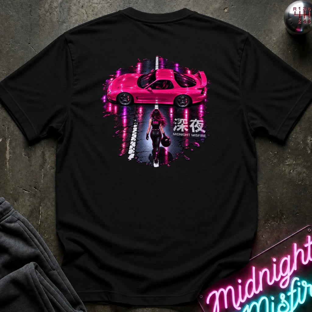 A black short-sleeve t-shirt laid flat on a dark textured surface, featuring a large graphic print on the back with a pink sports car, a woman walking, Japanese characters