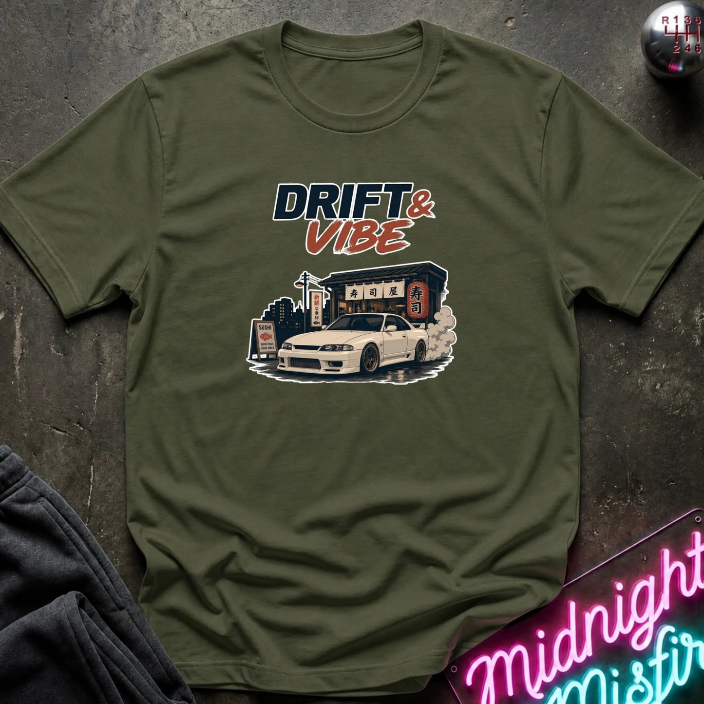 An olive green short-sleeve t-shirt with a 'DRIFT & VIBE' graphic print featuring a white sports car and a drift shop, laid flat on a dark textured surface with a neon sign and dark pants partially visible.