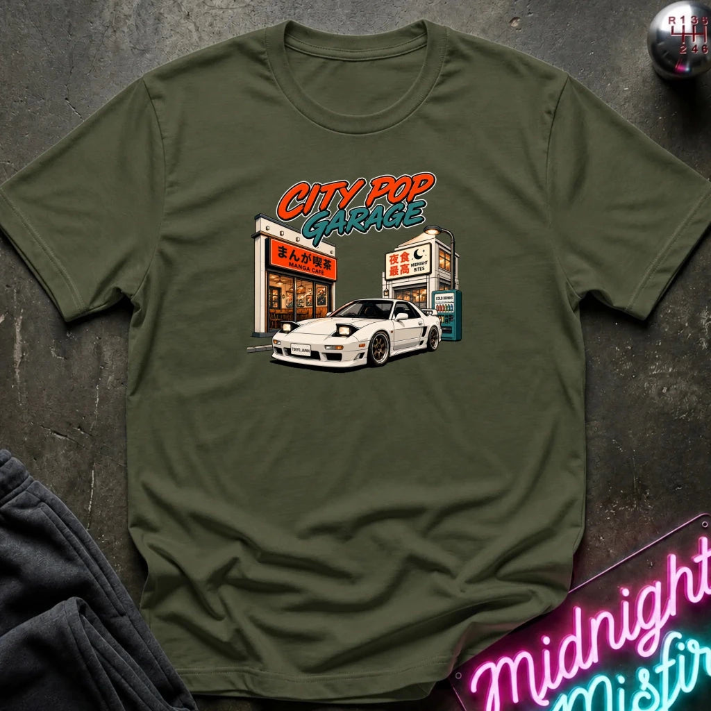 An olive green short-sleeve t-shirt laid flat on a dark textured surface, featuring a graphic print of a white sports car parked outside a garage with the text 'CITY POP GARAGE'.