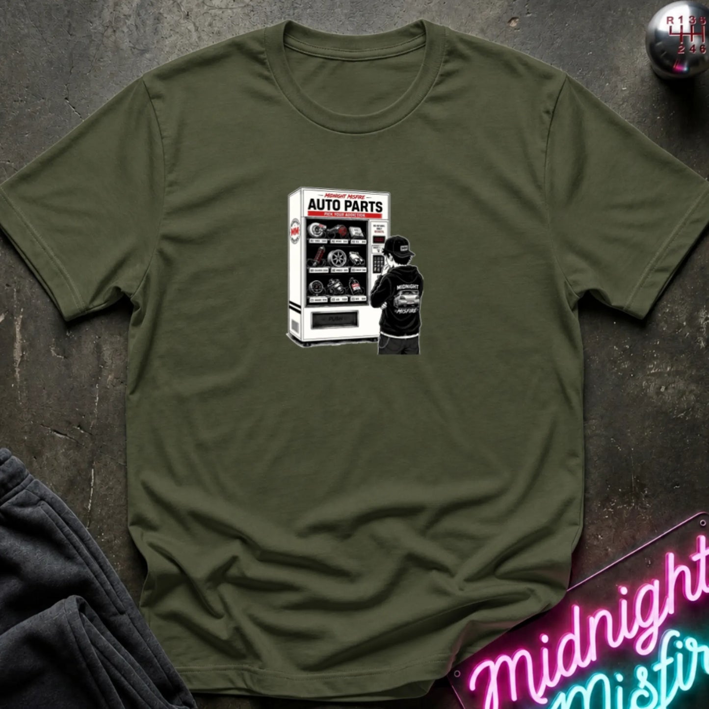 A olive green short-sleeve t-shirt laid flat on a dark textured surface, featuring a graphic print of a vending machine and owner deciding.