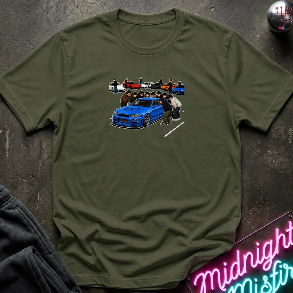 olive short-sleeve crewneck t-shirt laid flat on a dark textured surface. The center of the shirt features a graphic print of a blue sports car with a group of people standing behind it.