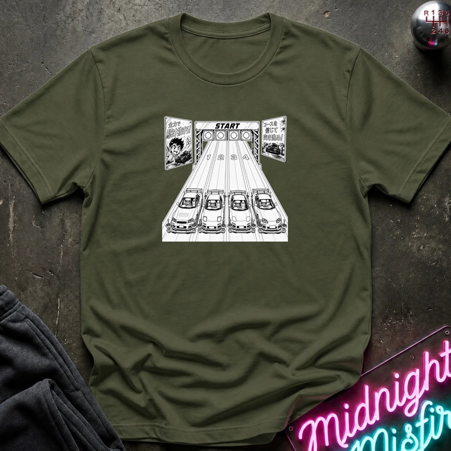 An olive short-sleeve t-shirt laid flat on a dark textured surface, featuring a black-and-white graphic of 4 cars lined up at a racing on a track.