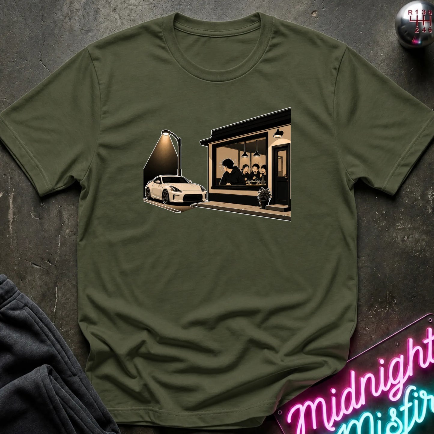 An olive green short-sleeve t-shirt laid flat on a dark textured surface, featuring a graphic print of a white sports car parked outside a dimly lit cafe with a neon sign in the foreground.