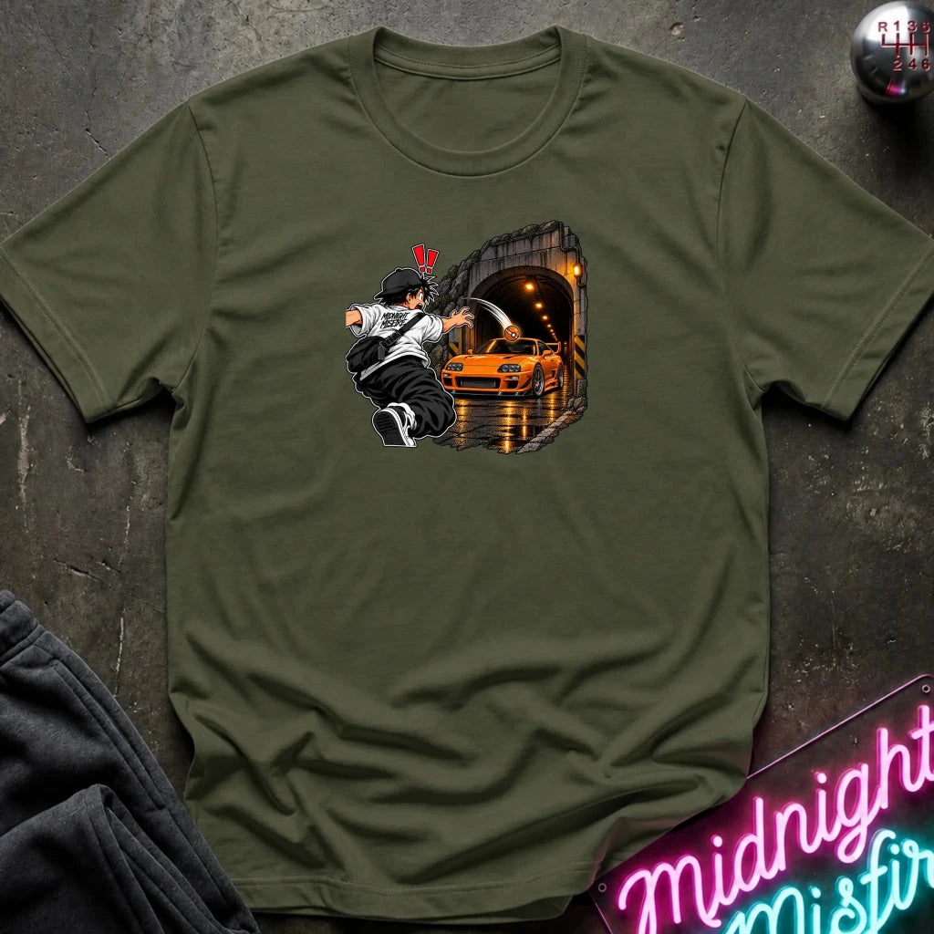 An olive green short-sleeve crewneck t-shirt laid flat on a dark textured surface. The center of the shirt features a graphic print of an anime character in a black and white outfit with a hat, next to a sports car 
