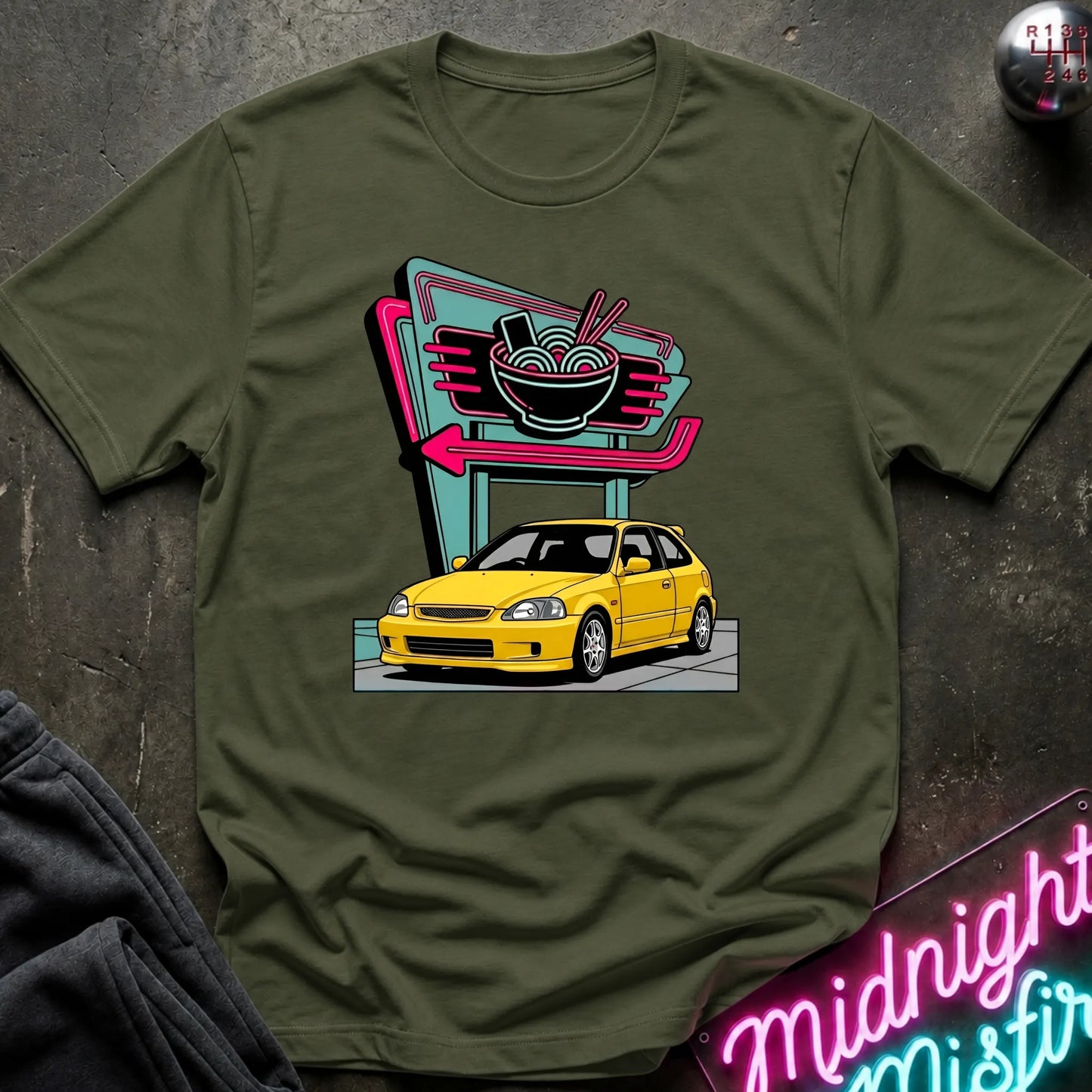  An olive  green short-sleeve graphic t-shirt laid flat on a dark textured surface. The shirt features a colorful retro-style print of a yellow car parked in front of a neon ramen shop sign.