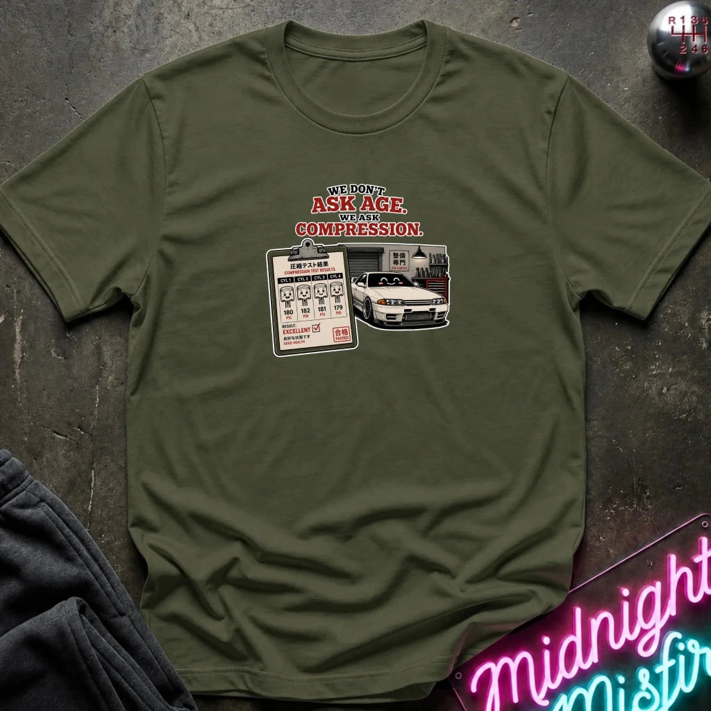An olive green short-sleeve graphic t-shirt laid flat on a dark textured surface. The graphic features the text 'WE DON'T ASK AGE, WE ASK COMPRESSION.