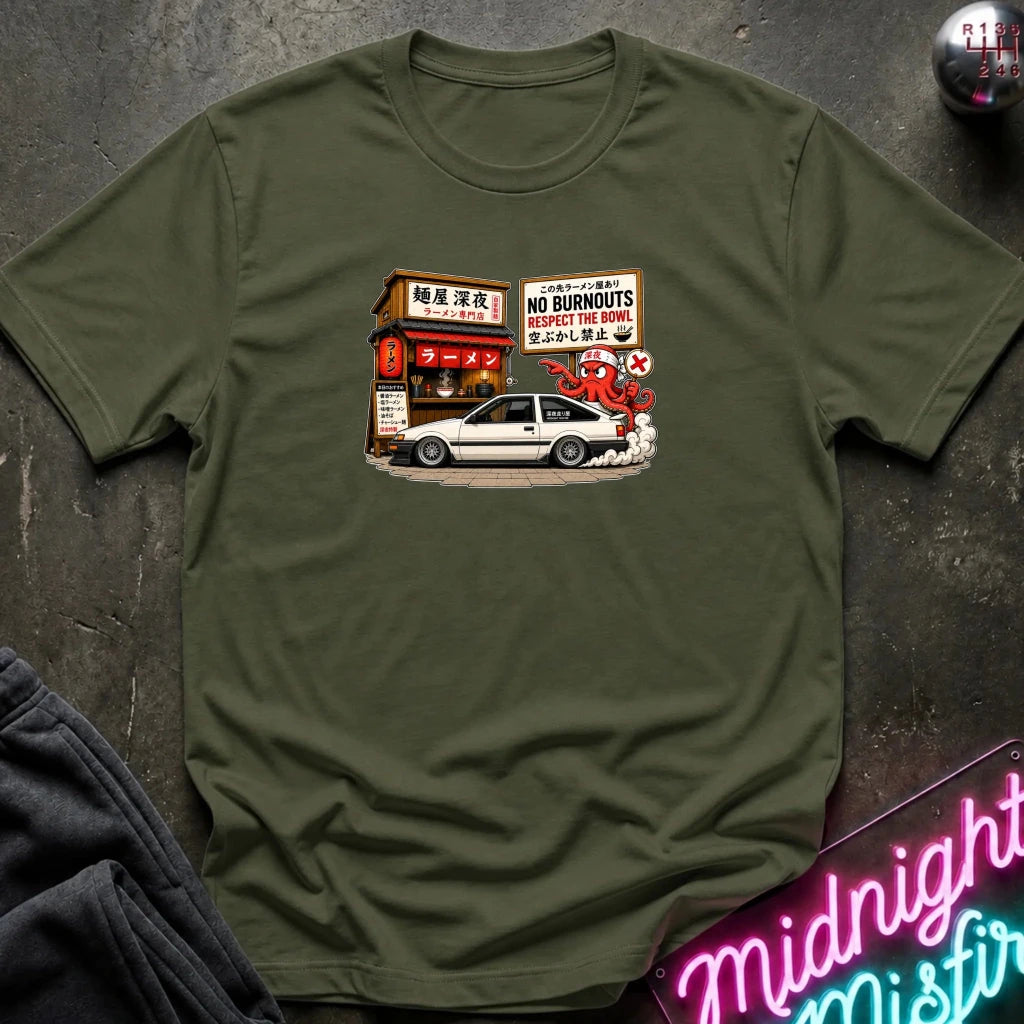 An olive green crewneck t-shirt featuring a graphic print of a white sports car parked in front of a Japanese restaurant with a 'No Burnouts' sign, styled flat on a dark textured surface with a neon sign and sweatpants partially visible.