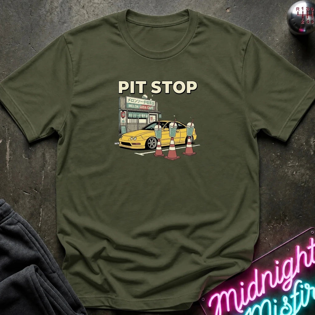 An olive green short-sleeve t-shirt with a graphic print of a yellow sports car at a pit stop, labeled 'PIT STOP' in beige text. The shirt is laid flat on a dark textured surface with a neon sign and dark pants partially visible in the corners.