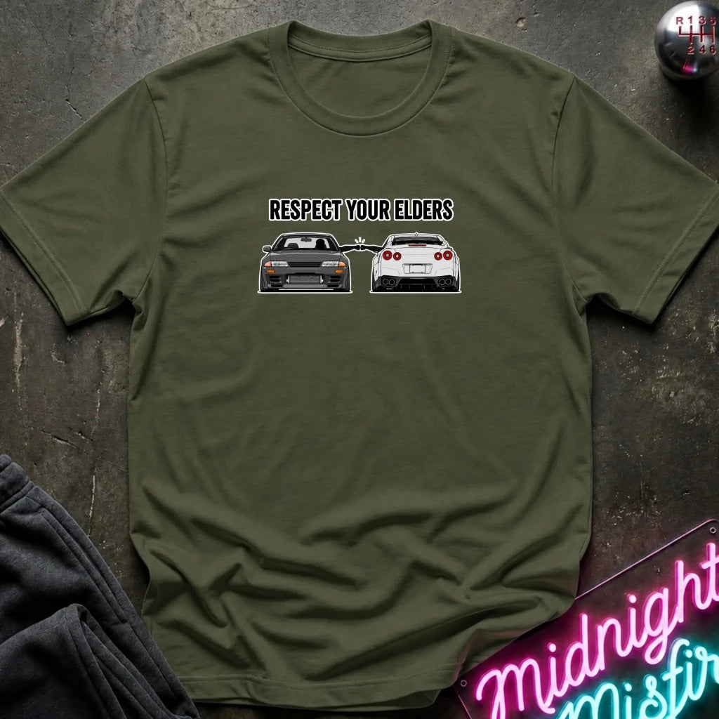 An olive green short-sleeve t-shirt laid flat on a dark textured surface. The shirt features a graphic print with the text 'RESPECT YOUR ELDERS' above an illustration of a black classic car facing a white modern sports car.