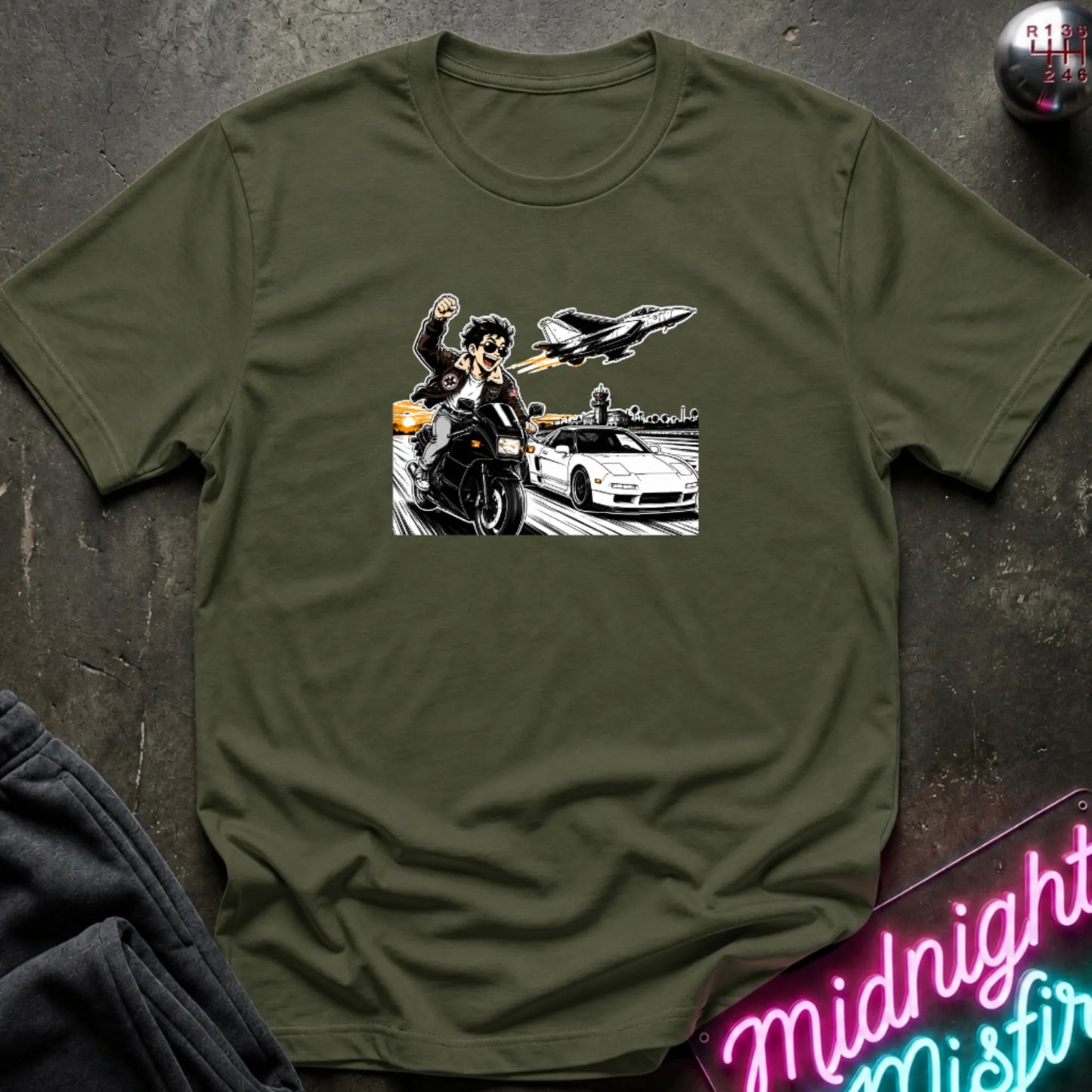 A ollive green short-sleeve crewneck t-shirt with a graphic print of a motorcycle racer, a sports car, and a jet plane, laid flat on a dark textured background with a neon sign and sweatpants partially visible.