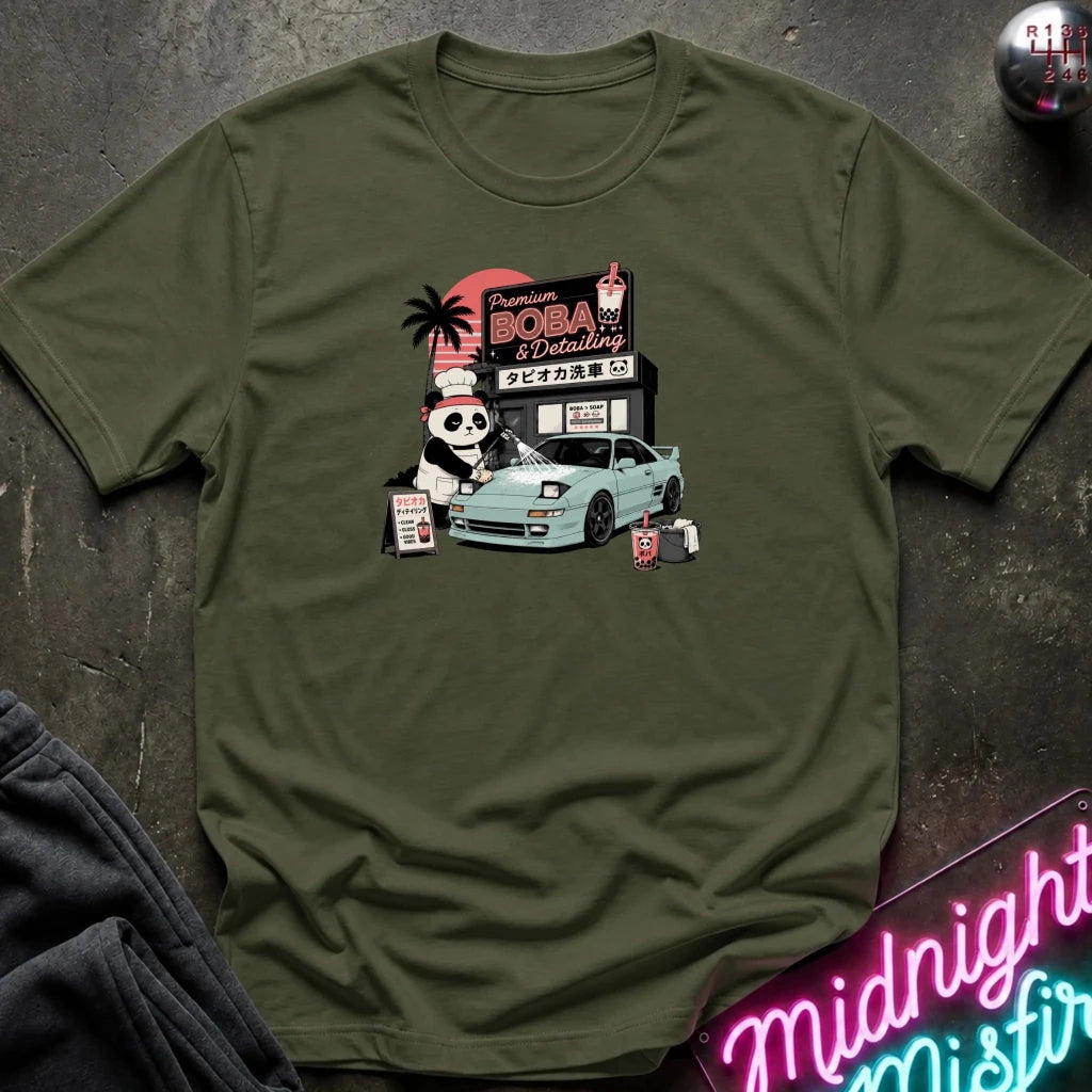 olive short-sleeve graphic t-shirt laid flat on a dark textured surface. The graphic features a cartoon panda washinga light blue car