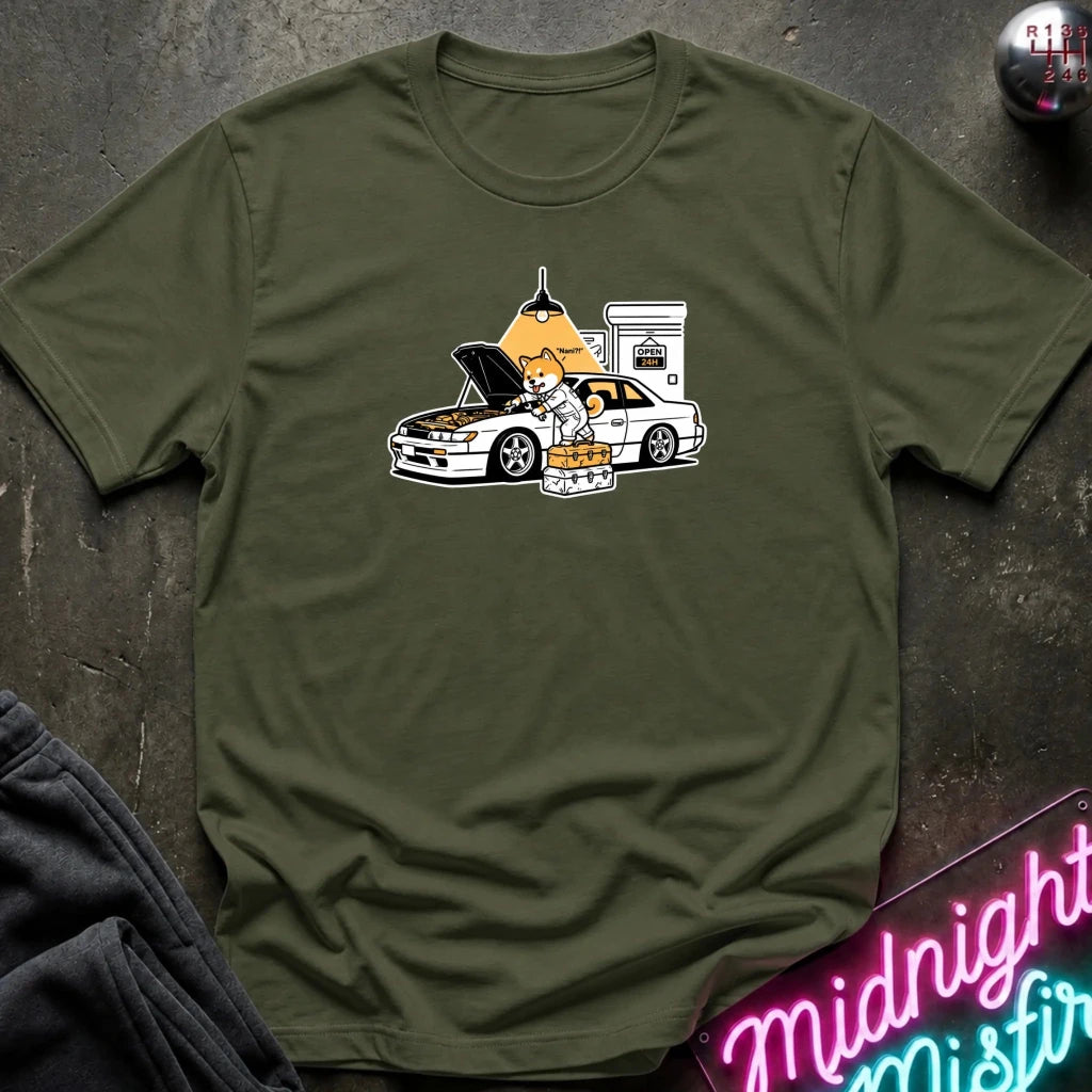 olive graphic t-shirt laid flat on a dark textured surface. The graphic features a dog fixing a car.