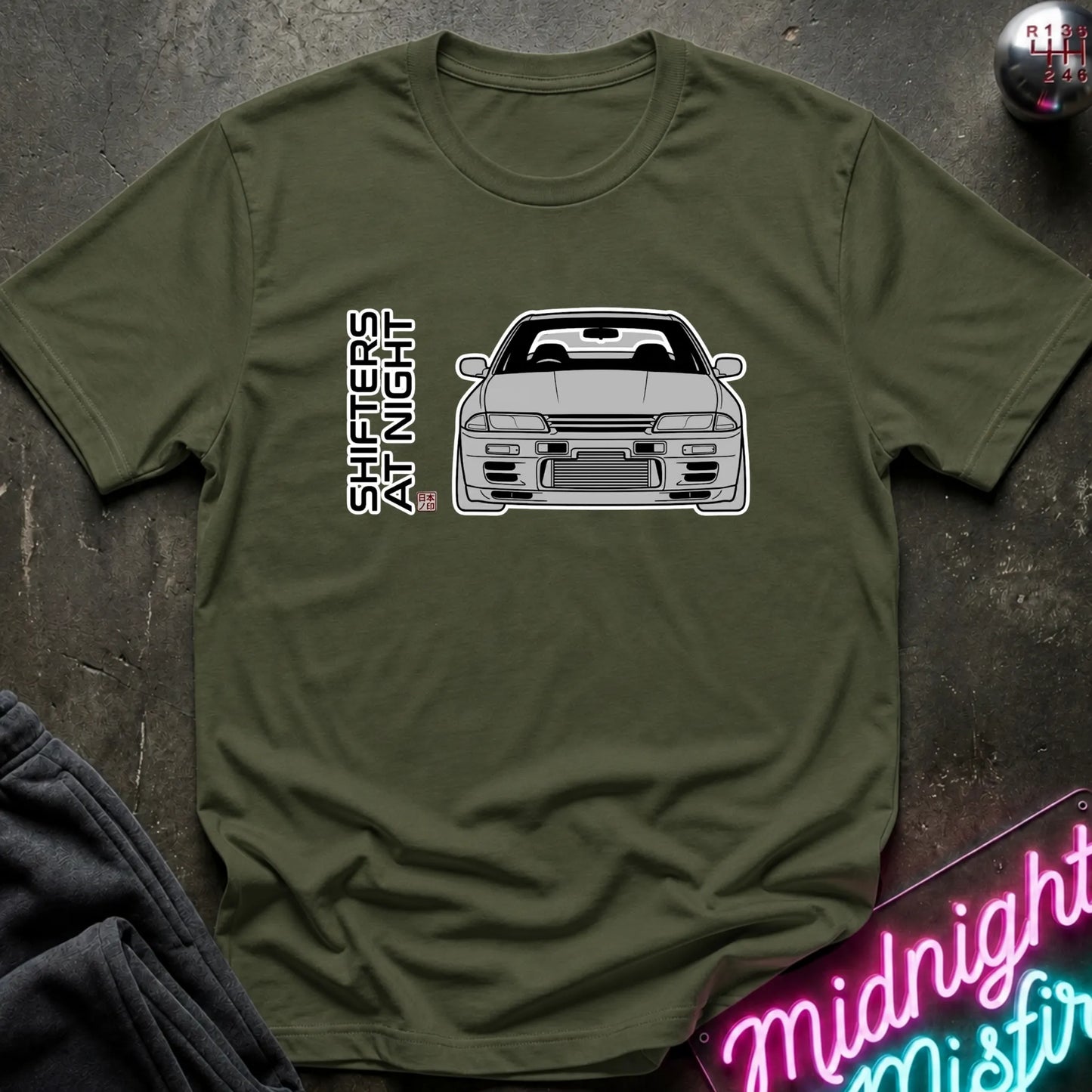 An olive green short-sleeve t-shirt with a graphic of a white sports car and the vertical text 'SHIFTERS AT NIGHT' on the chest, displayed flat on a dark textured surface with a neon sign and dark pants in the background.