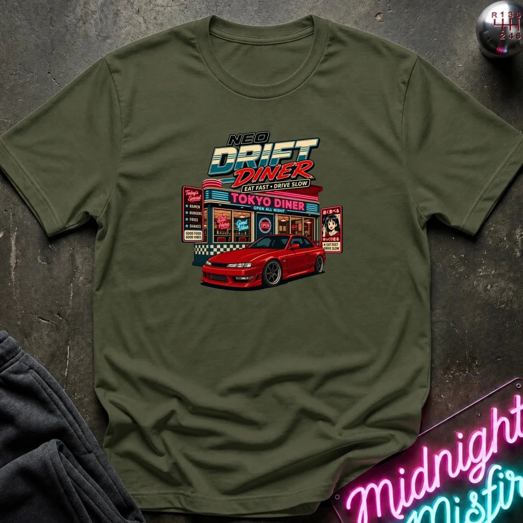 An olive green crewneck t-shirt featuring a colorful graphic print of a 'NBA Drift Diner' with a red sports car, laid flat on a dark textured surface with a neon sign and dark pants partially visible.