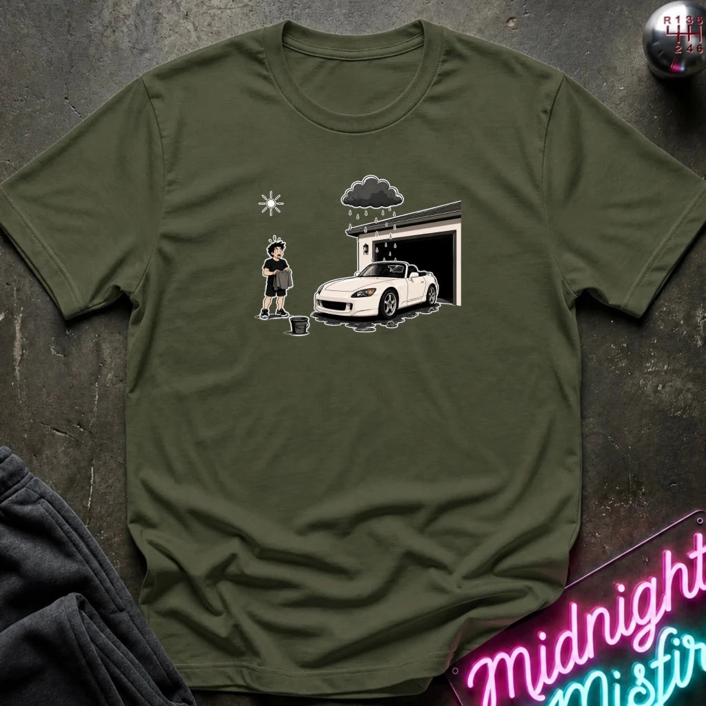 An olive green short-sleeve t-shirt laid flat on a dark textured surface, featuring a cartoon graphic of a person washing a white sports car in front of a garage. 