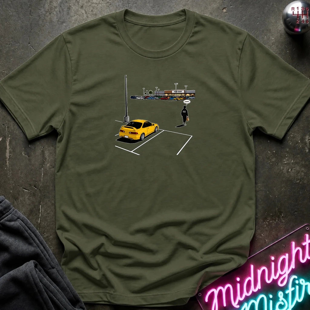 An olive green short-sleeve t-shirt laid flat on a dark textured surface, featuring a small graphic print of a yellow car, a parking lot, and a lone figure. 