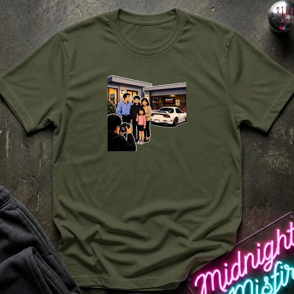An olive green short-sleeve crewneck t-shirt laid flat on a dark textured surface, featuring a rectangular graphic print of a family standing outside a building taking a photo.